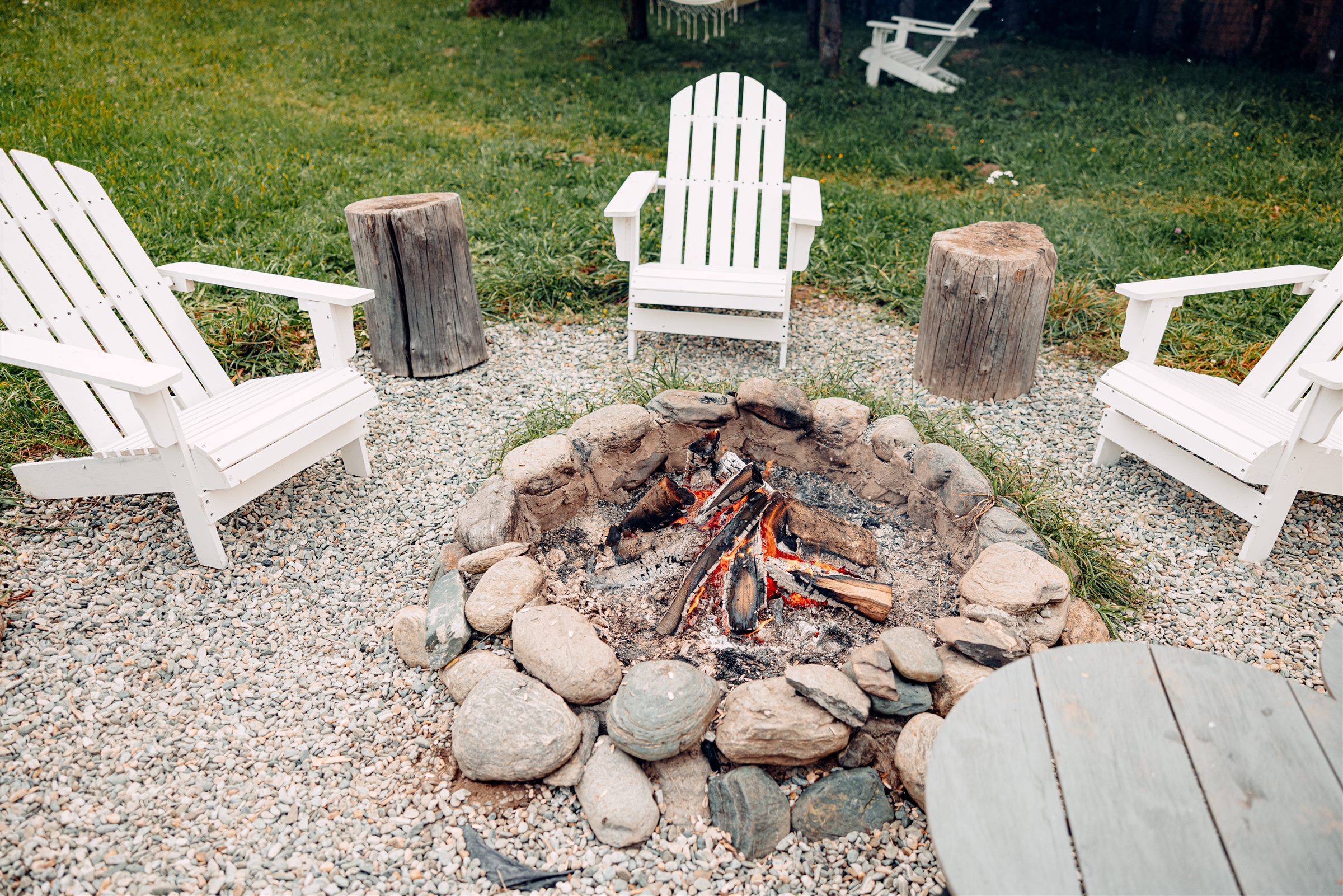 Stone fire pit ringed by white Adirondack chairs on a gravel patio
